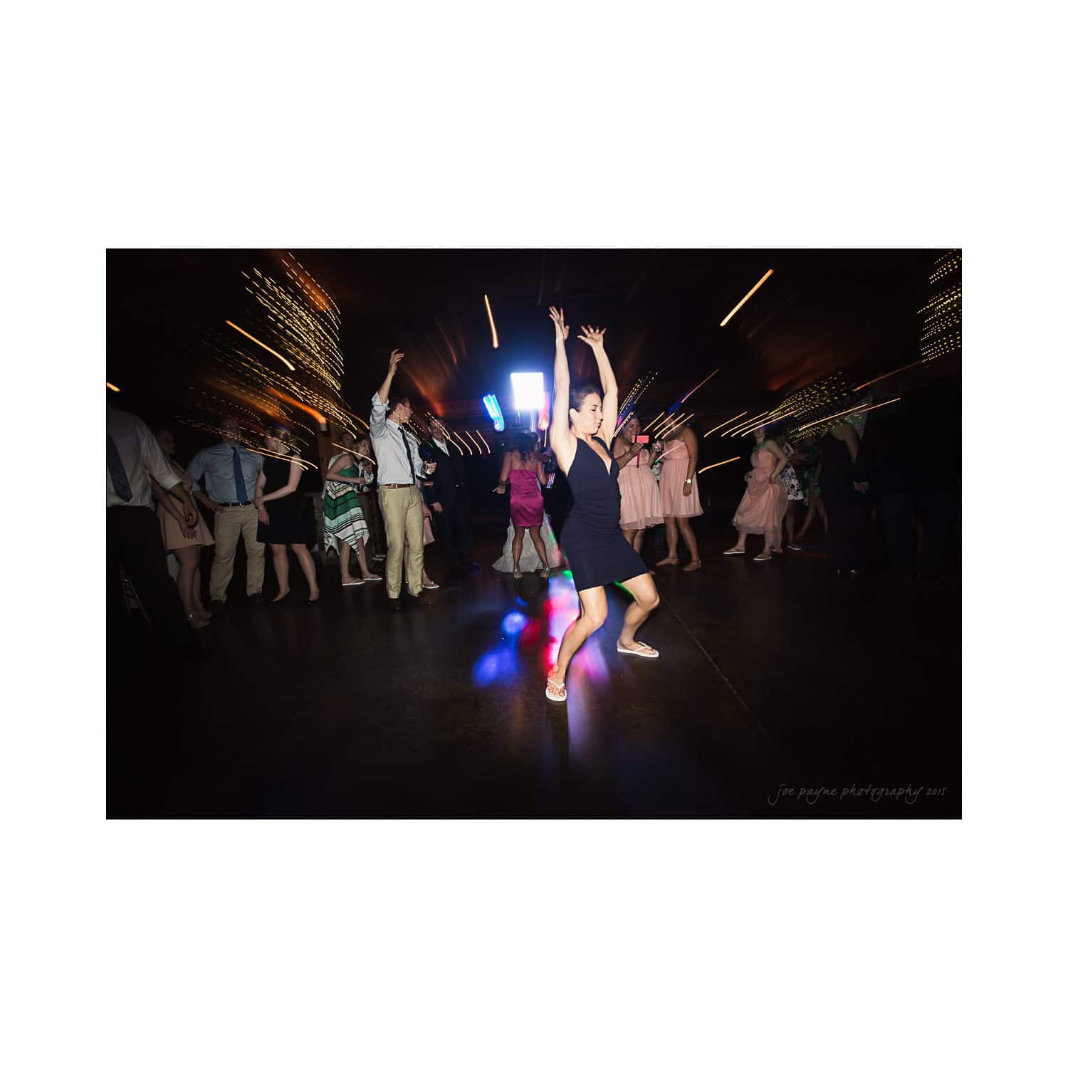 A lively dance floor scene with colorful lights reflecting off the floor. The central focus is a person in a black dress with hands raised, surrounded by people dancing energetically. The atmosphere is vibrant and festive.
