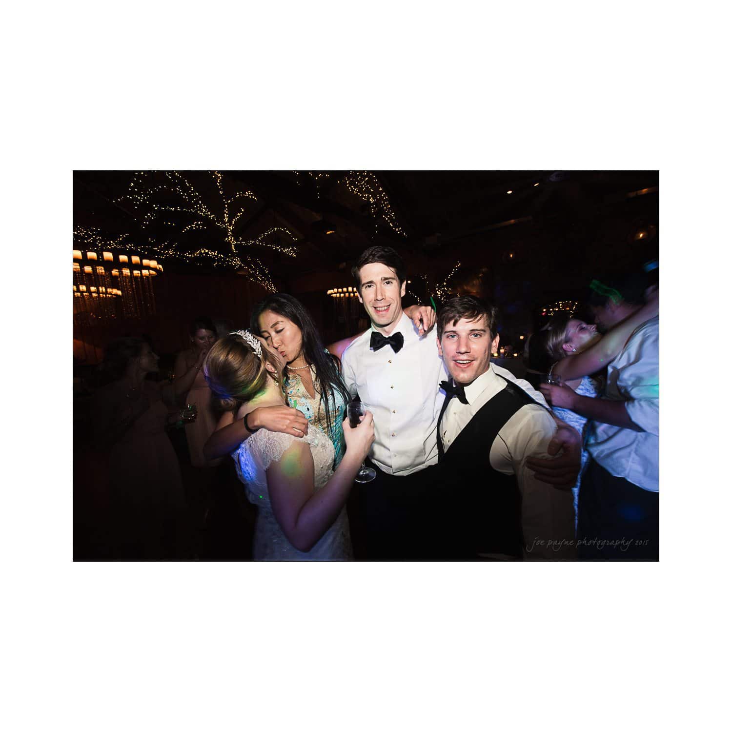 A group of four people dressed in formal wear, posing together at a party. The setting features dim lighting with decorative lights in the background, creating a festive atmosphere.