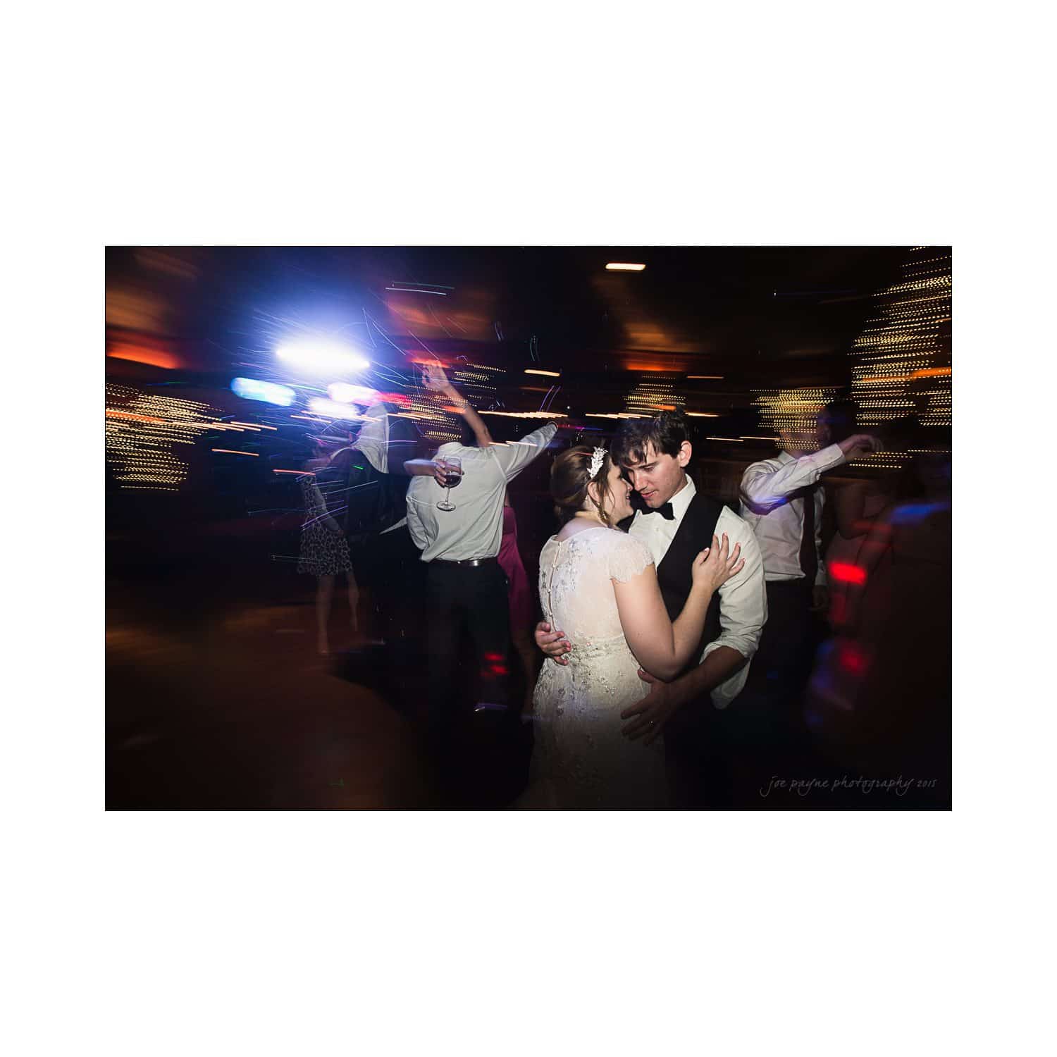 A couple dances closely at a dimly lit event with colorful lights. Blurred figures dance in the background, creating a lively atmosphere. The scene suggests a joyful celebration or wedding reception.