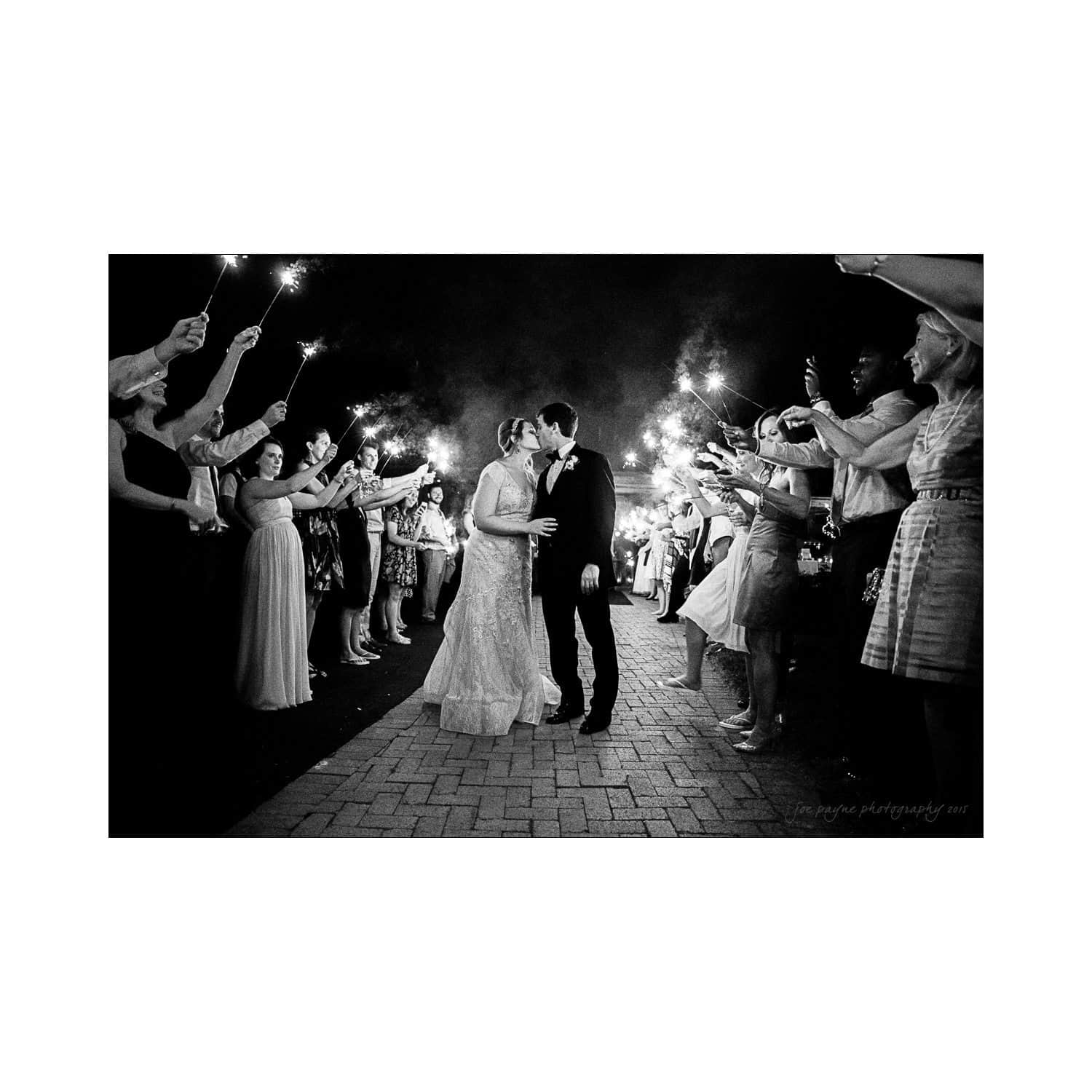 A bride and groom share a kiss on a brick pathway, surrounded by guests holding sparklers. The scene is illuminated by the sparklers, capturing the joyous atmosphere of the celebration.