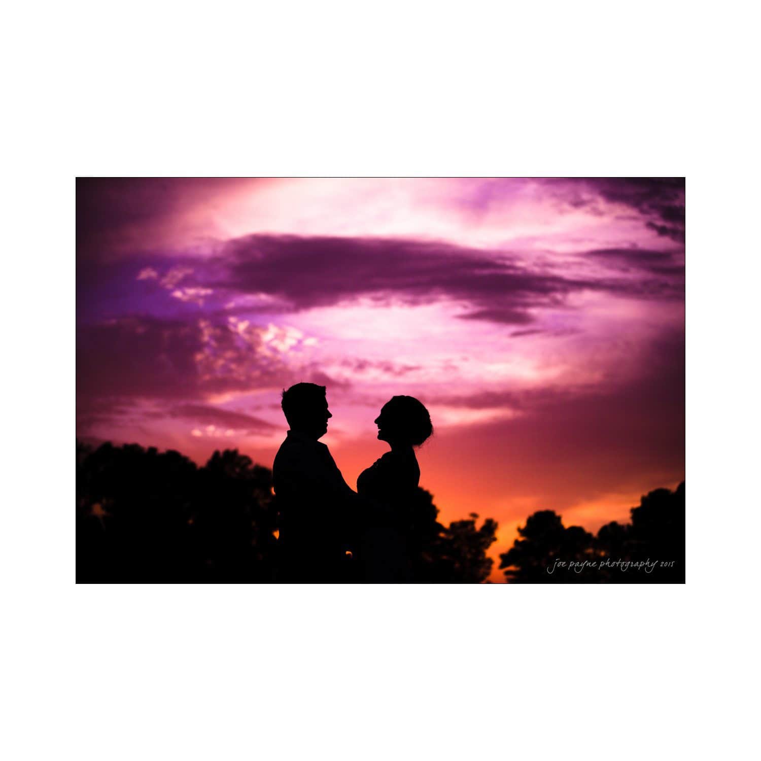 chapel hill wedding photography - nicole & wayne Silhouette of a couple standing close together against a vibrant, multicolored sunset sky with shades of purple, pink, and orange. Trees are visible in the background.