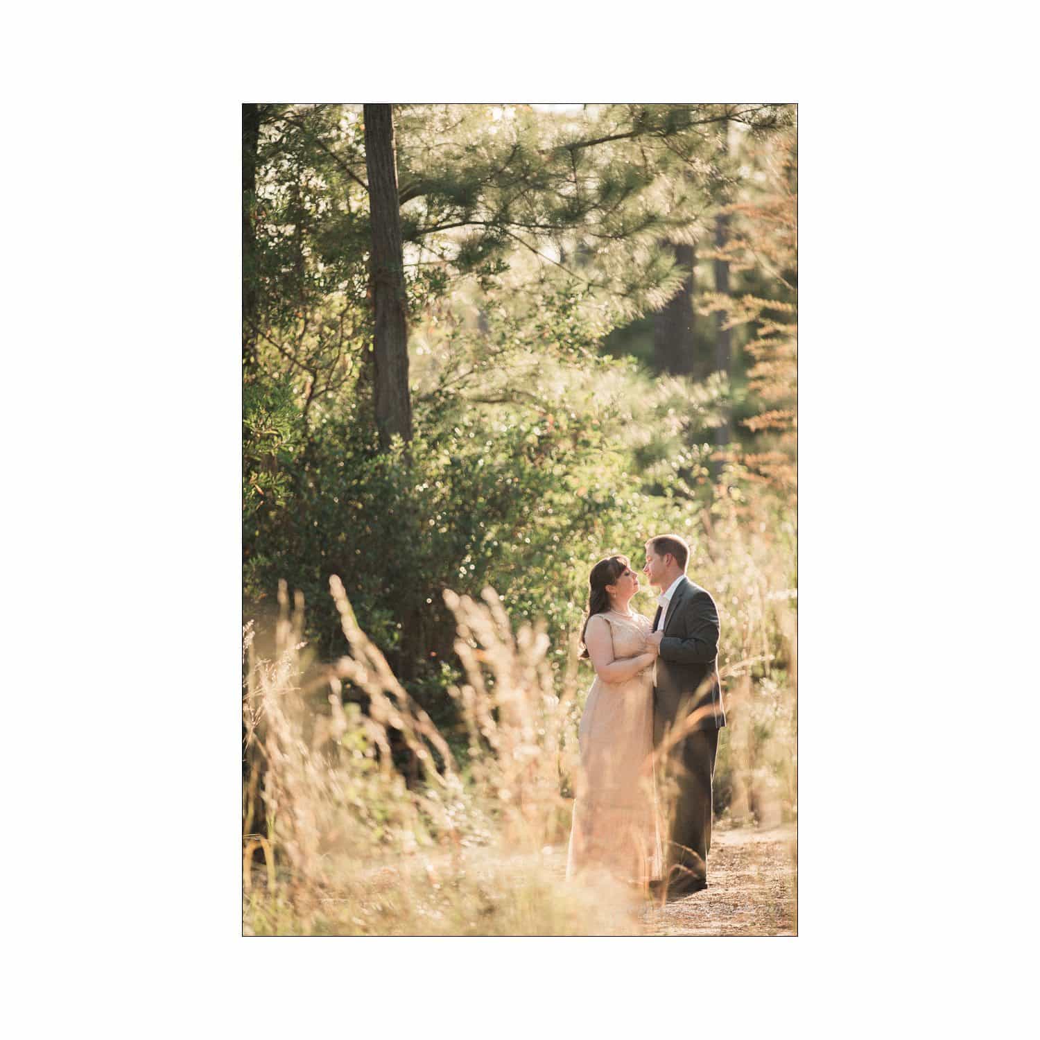 A couple stands closely together in an outdoor setting bathed in warm sunlight. Tall grasses and trees surround them, creating a romantic and serene atmosphere.
