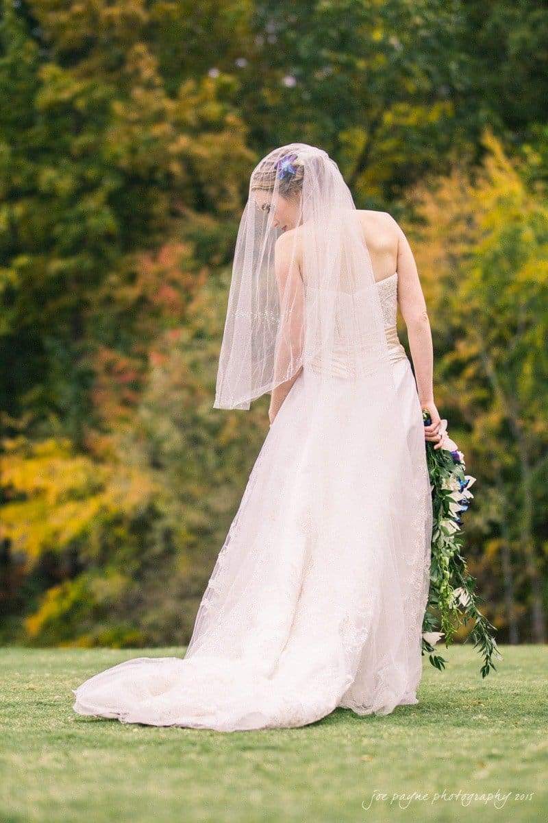 charlotte wedding photographers - ashley & matt A bride stands outdoors, facing away, in a strapless white gown and veil. She holds a bouquet of purple and white flowers. The background is a blurred view of lush green and autumn-colored trees.
