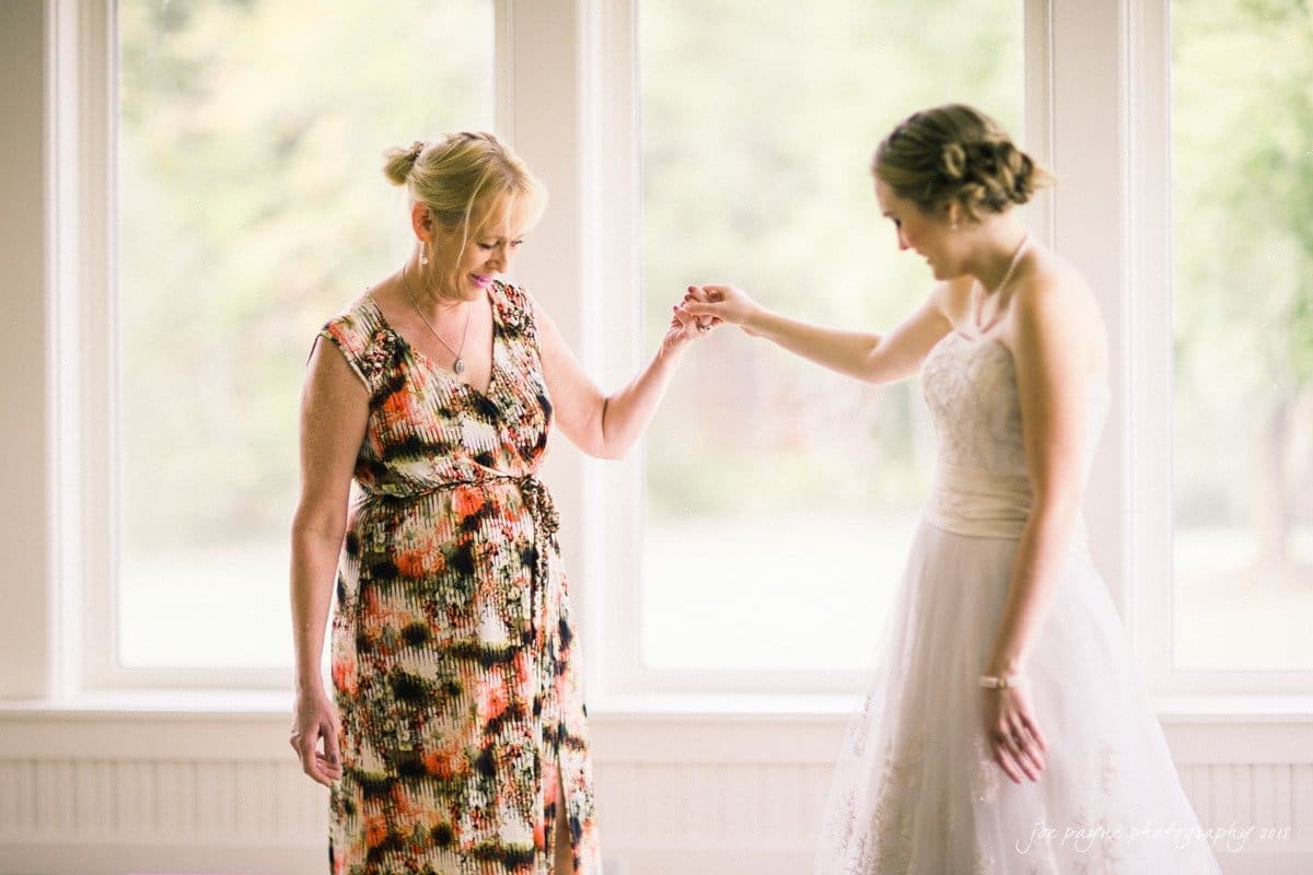 charlotte wedding photographers - ashley & matt A bride in a white dress and an older woman in a floral dress hold hands and smile at each other indoors, in front of large windows. The background is softly blurred, highlighting the warm moment between them.