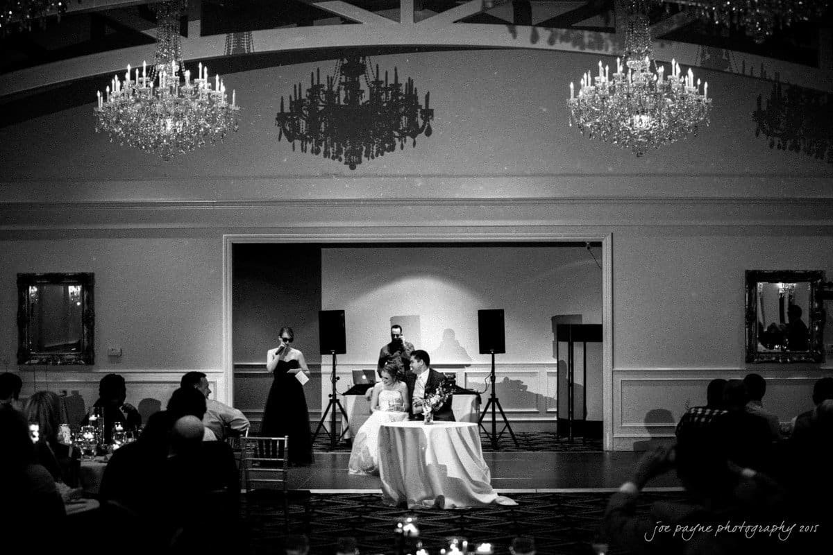 charlotte wedding photographers - ashley & matt A black and white photo of a wedding reception with a couple seated at a table in the center. A woman stands with a microphone, and guests are seated around them. Chandeliers hang from the ceiling, adding an elegant touch to the scene.