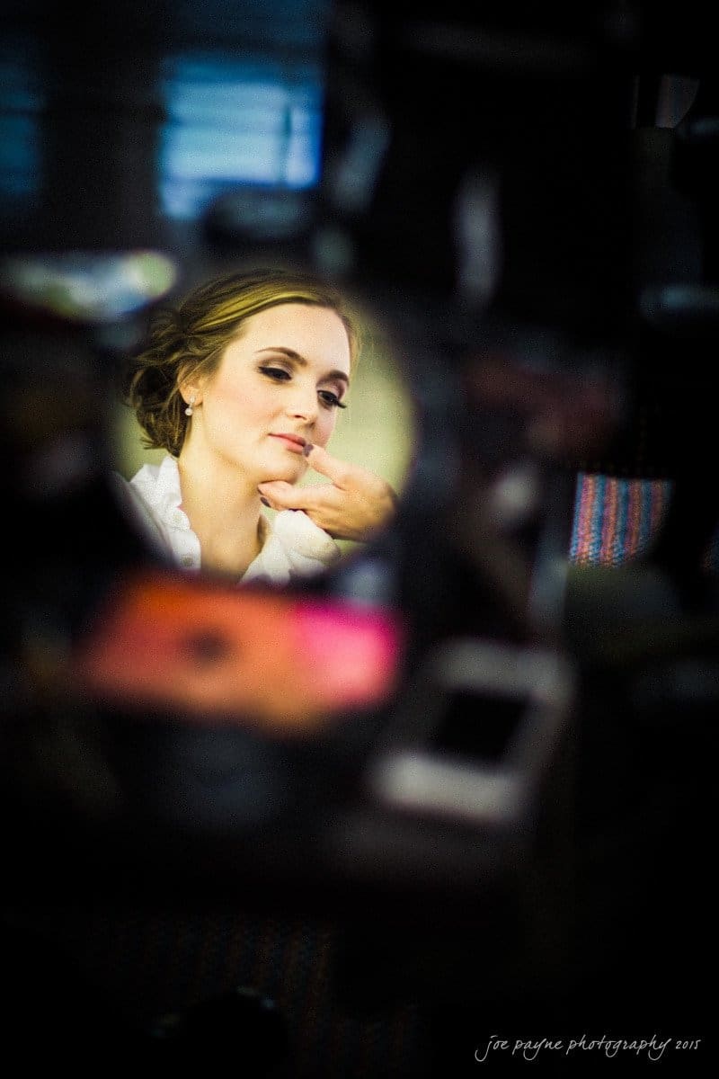 charlotte wedding photographers - ashley & matt A woman is having her makeup applied, reflected in a round mirror. She has her hand under her chin and is surrounded by blurred objects.