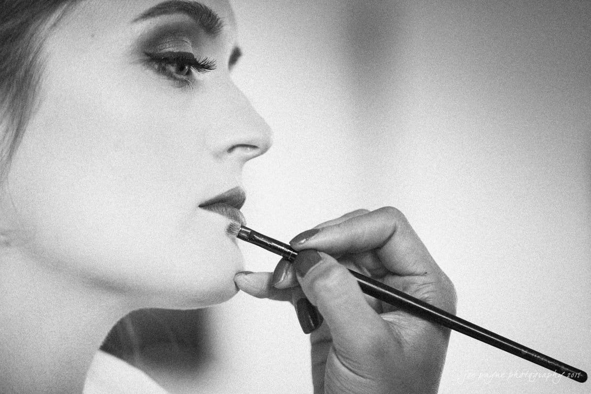 charlotte wedding photographers - ashley & matt Black and white close-up of a person with eyes closed, receiving makeup on their lips. A hand holds a brush applying the makeup.