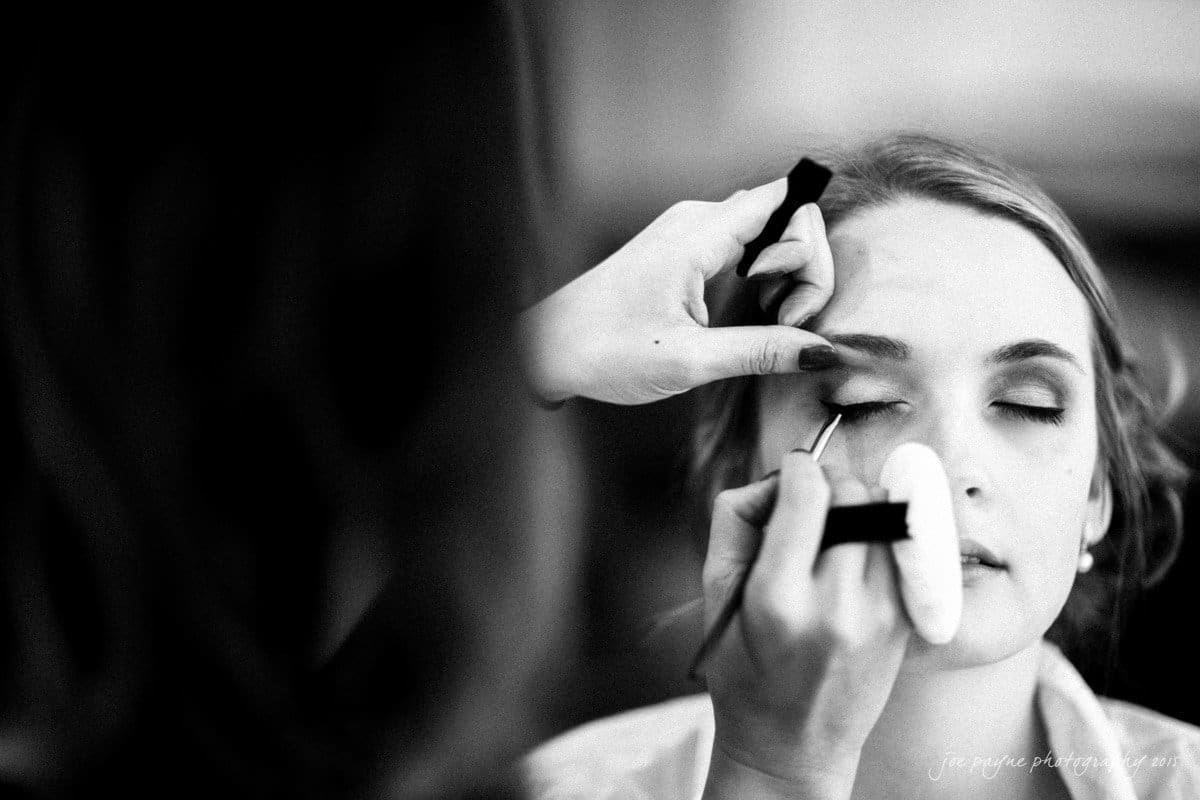 charlotte wedding photographers - ashley & matt Black and white image of a person with closed eyes having makeup applied by another persons hands. The makeup artist uses a brush on the eyelid, while holding a compact. The scene is focused on the face, conveying a sense of calm and concentration.