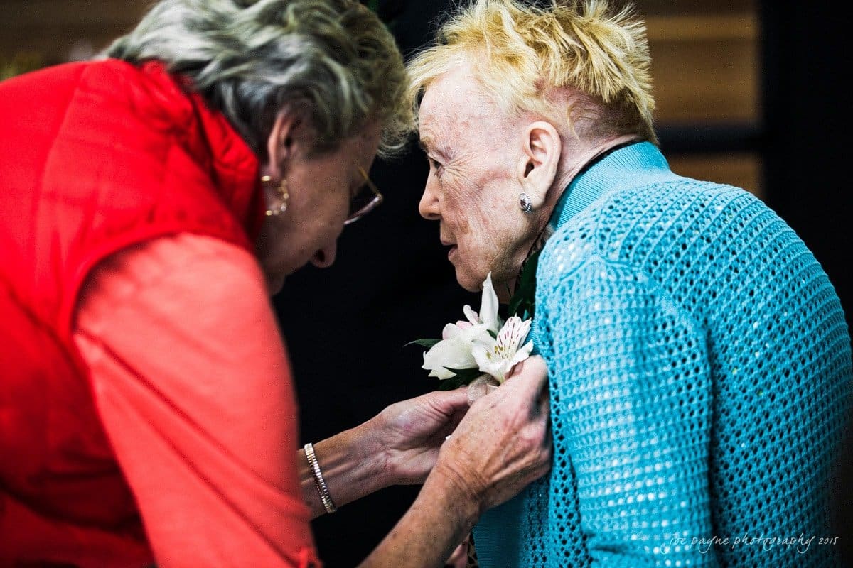 charlotte wedding photographers - ashley & matt An elderly woman in a blue knit sweater leans forward as another woman, wearing a red vest, adjusts a floral corsage on her chest. They appear engaged in a gentle exchange in an indoor setting.