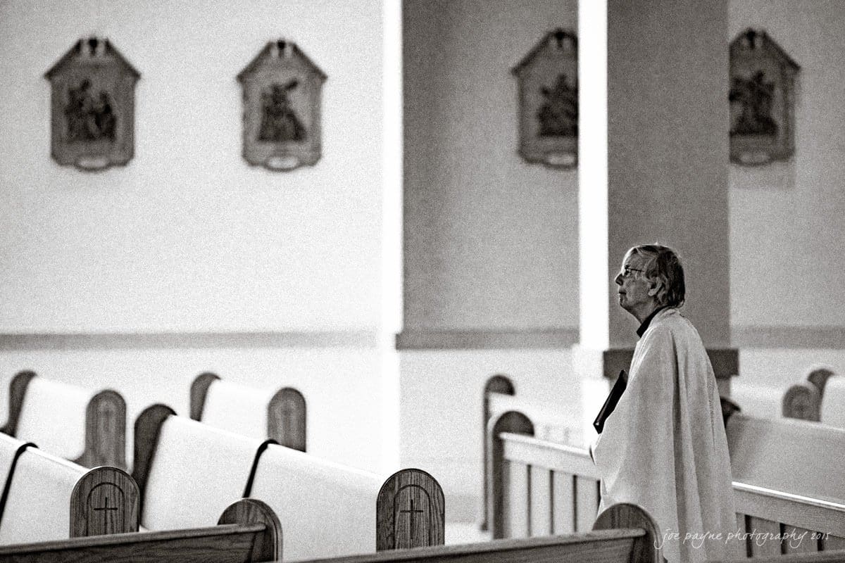 charlotte wedding photographers - ashley & matt A person stands alone in a church, wearing a coat, with empty pews surrounding them. The atmosphere is calm and reflective, with religious plaques visible on the walls.