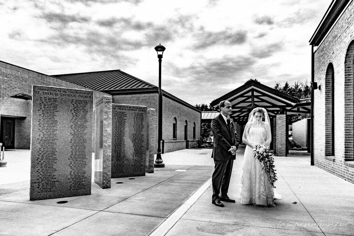 charlotte wedding photographers - ashley & matt A bride and groom stand together in front of large engraved stone plaques outside a building with a covered walkway. The sky is cloudy, and the scene is in black and white. The bride holds a bouquet, and both appear solemn.