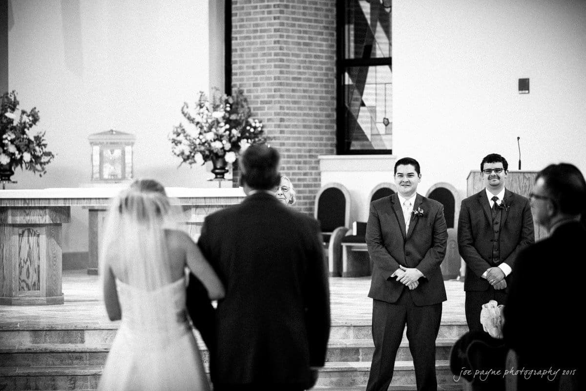 charlotte wedding photographers - ashley & matt A bride, escorted by an older man, walks down the aisle toward a groom in a suit and groomsmen in a church. The groom smiles as he watches her approach. Floral arrangements decorate the altar in the background.