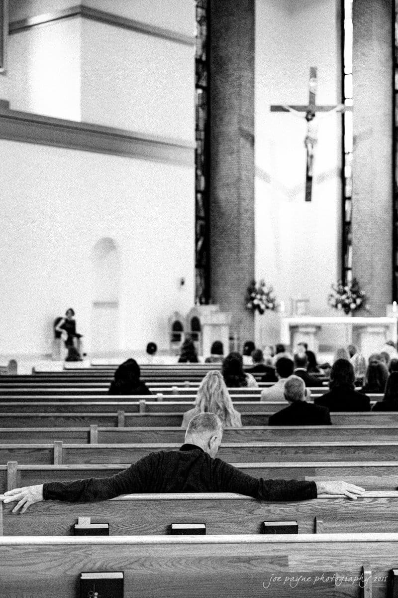 charlotte wedding photographers - ashley & matt Black and white photo of a church interior during a service. A person sits in a pew near the back with arms stretched along the backrest. The altar is visible in the distance with a crucifix and floral arrangements.