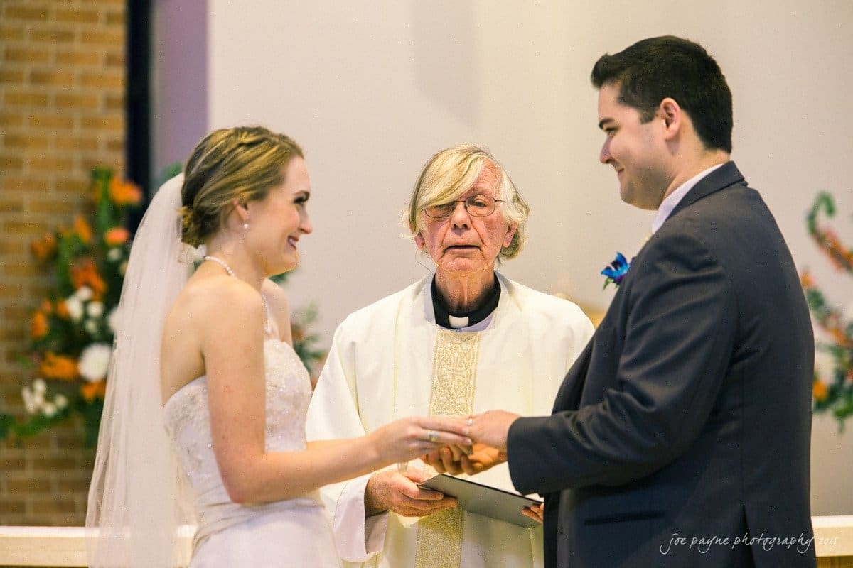 charlotte wedding photographers - ashley & matt A bride and groom stand facing each other, holding hands, in a church setting. An officiant in a white robe stands between them, smiling as he reads from a book. Floral decorations are visible in the background.
