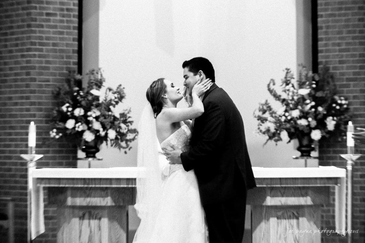 charlotte wedding photographers - ashley & matt A bride and groom share a kiss in a wedding ceremony. They stand at the altar, surrounded by floral arrangements and lit candles. The setting is elegant and formal, with a focus on the couples embrace.
