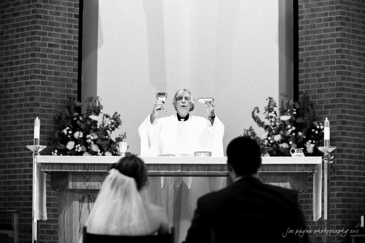 charlotte wedding photographers - ashley & matt A priest stands at an altar, holding up a chalice and bread. Two people, possibly a couple, sit facing the altar. The setting appears to be a church with floral arrangements and candles on the altar. The photo is in black and white.