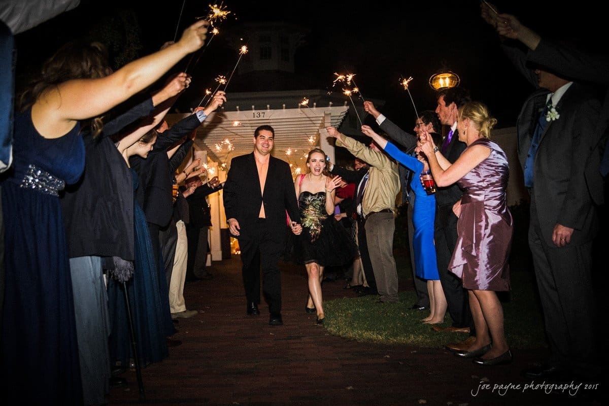 charlotte wedding photographers - ashley & matt A couple walks hand in hand through a sparkler-lit path at night, surrounded by cheering guests. The man wears a black outfit, and the woman wears a black and gold dress, smiling joyfully. The scene is celebratory and festive.