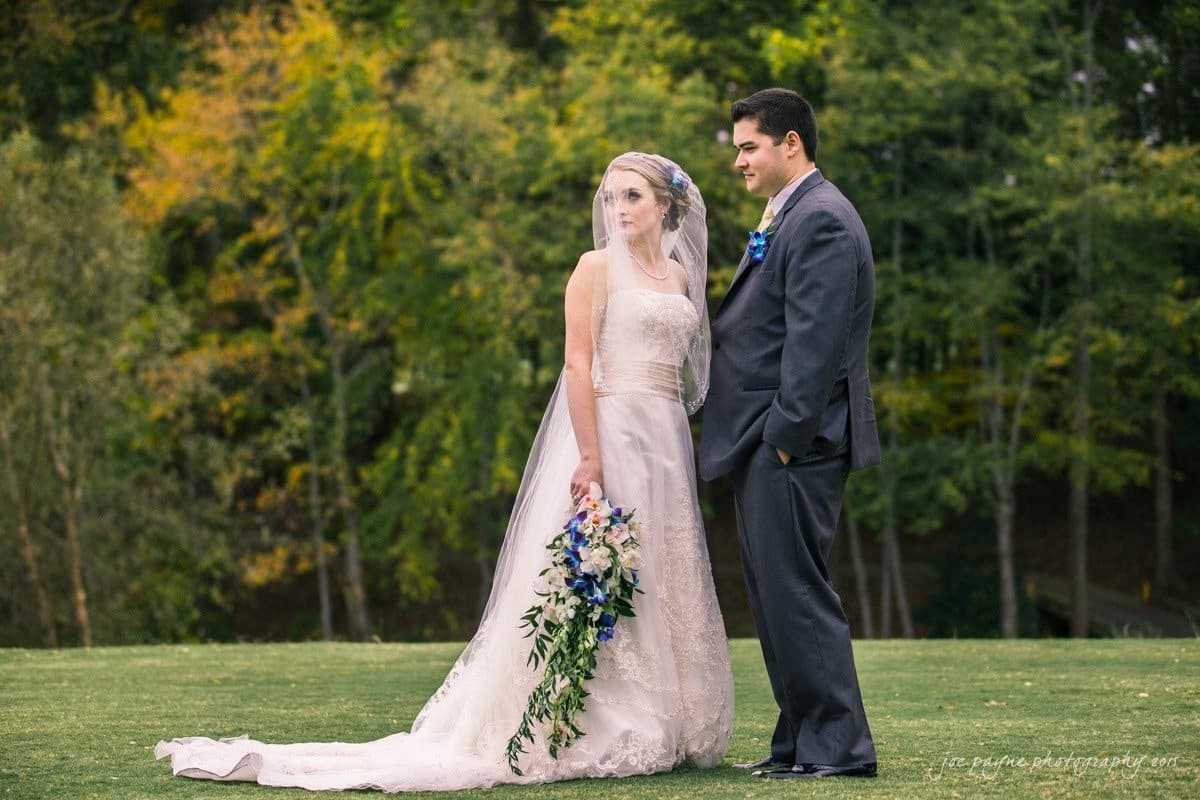 charlotte wedding photographers - ashley & matt A bride and groom stand together outdoors on a grassy area. The bride is wearing a strapless white gown and veil, holding a bouquet of white and blue flowers. The groom is in a dark suit. They are surrounded by lush green trees.