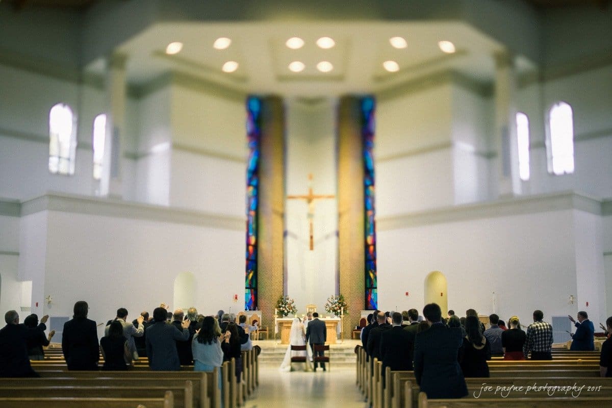 charlotte wedding photographers - ashley & matt A couple stands at the altar in a large, modern church with high ceilings. Guests are seated and standing, witnessing the ceremony. The space features stained glass windows and a prominent cross.