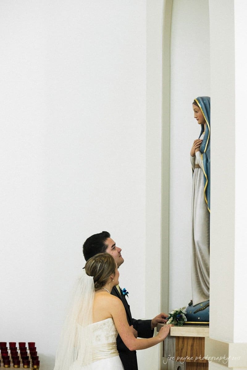 charlotte wedding photographers - ashley & matt A bride and groom stand in a church, looking at a statue of the Virgin Mary. The bride is wearing a white dress with a veil, and the groom is in a black suit. They appear to be placing flowers at the statues base.