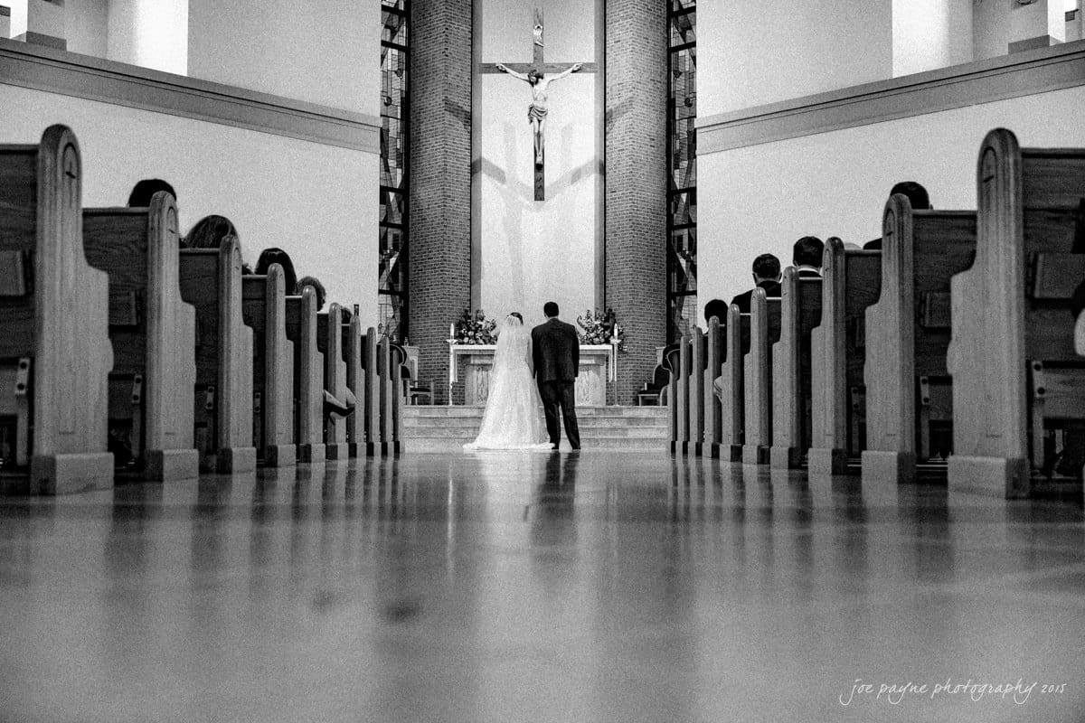 charlotte wedding photographers - ashley & matt A bride and groom stand at the altar in a church. The image is in black and white, showing pews lining both sides of the aisle leading to the couple and a cross on the wall behind them.
