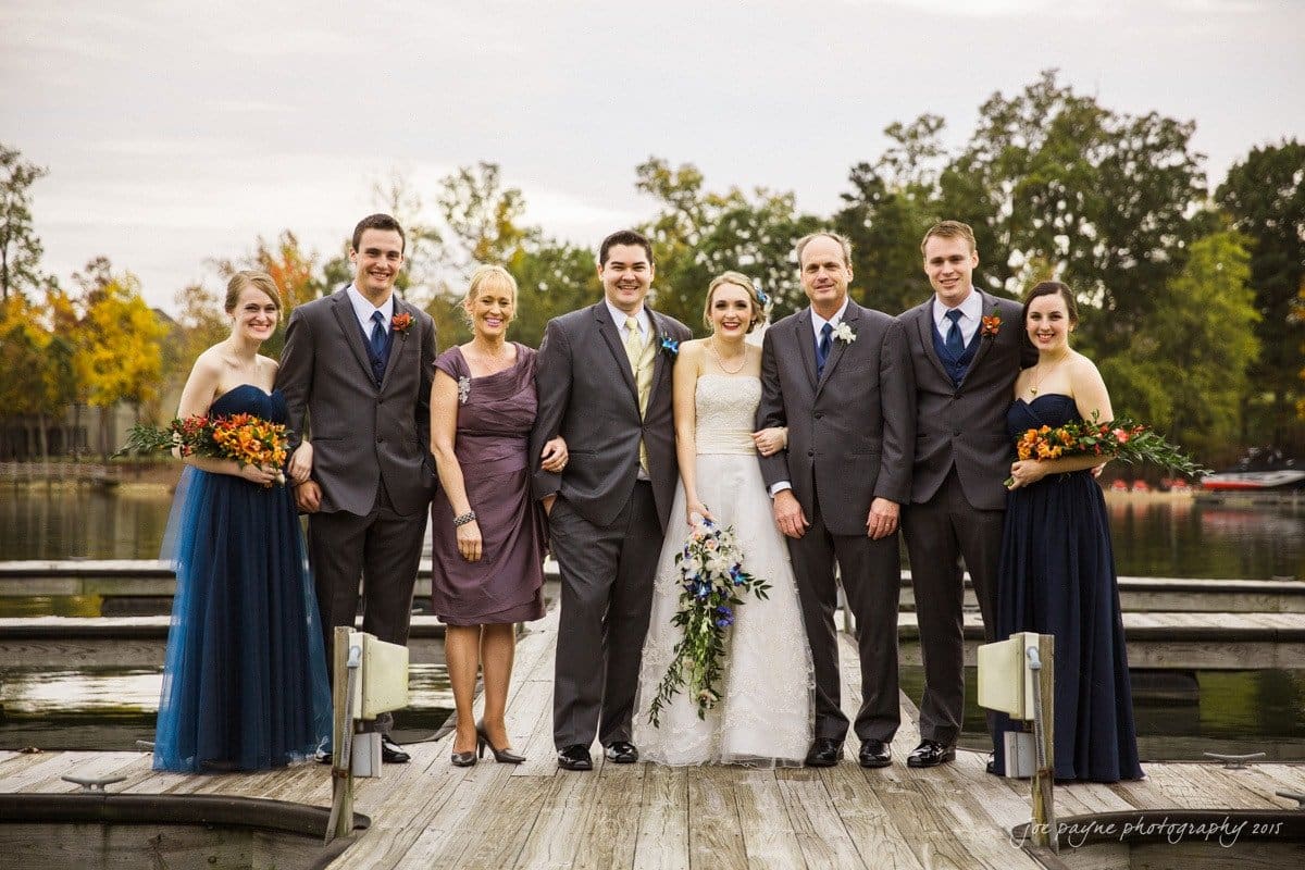 charlotte wedding photographers - ashley & matt A wedding group portrait on a lakeside dock. The bride and groom stand in the center, flanked by four bridesmaids in dark blue dresses holding bouquets, and two groomsmen in gray suits. Trees and a calm lake serve as the backdrop.
