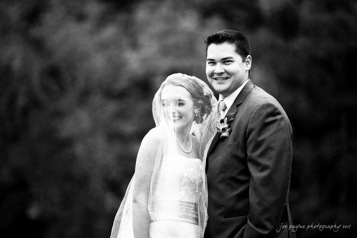 charlotte wedding photographers - ashley & matt A bride and groom smile happily in a black and white outdoor photo. The bride is wearing a veil, and the groom is in a suit with a boutonniere. Both appear joyful and content in a natural setting.