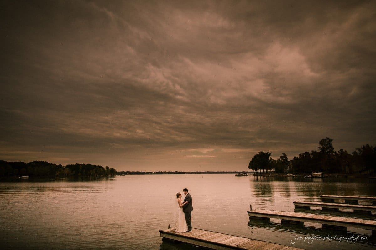 charlotte wedding photographers - ashley & matt A couple embraces on a wooden dock by a serene lake under a dramatic, cloud-filled sky. The water is calm, reflecting the muted tones of the overcast evening. Trees line the distant shore, enhancing the peaceful atmosphere.