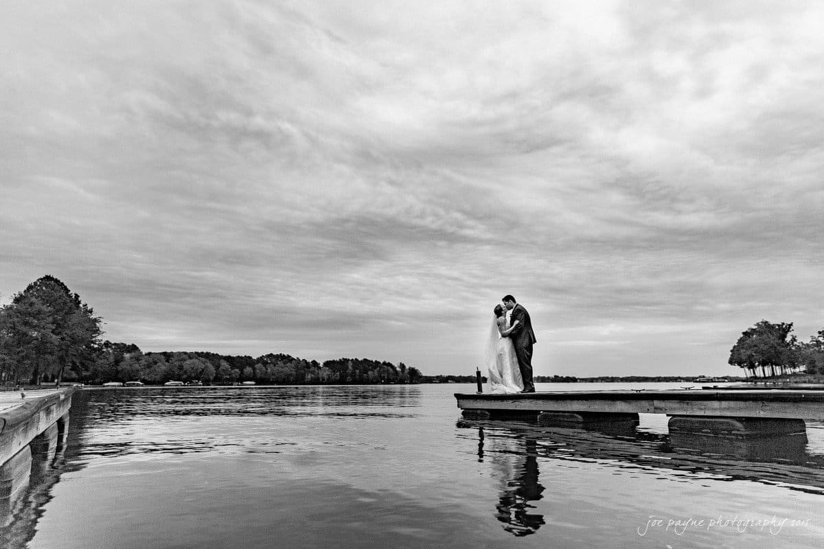 charlotte wedding photographers - ashley & matt A bride and groom stand on a wooden dock, embracing under a cloudy sky, with a tranquil lake surrounding them. Trees are visible in the distance, reflecting in the water, creating a peaceful and romantic scene.