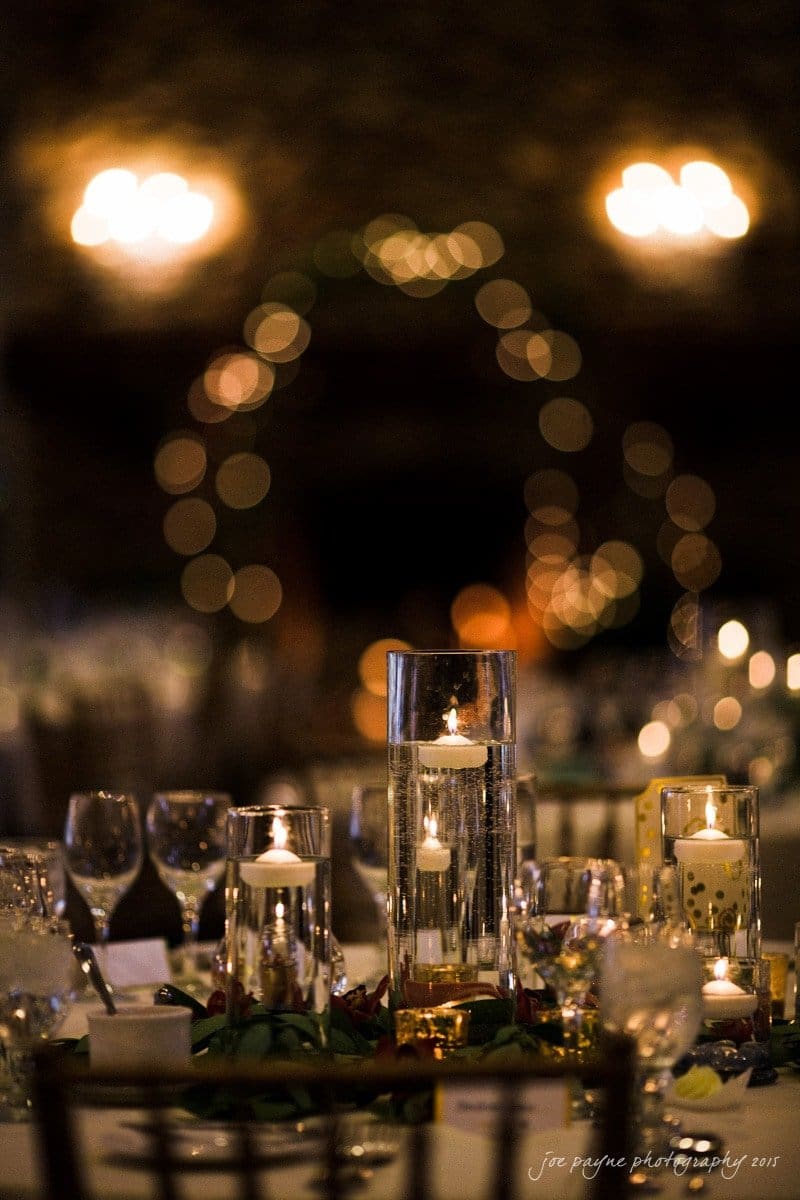 charlotte wedding photographers - ashley & matt A dimly lit table setting with floating candles in tall glass vases, surrounded by wine glasses and plates. In the background, blurred decorative lights form an arch, creating a warm, elegant atmosphere.