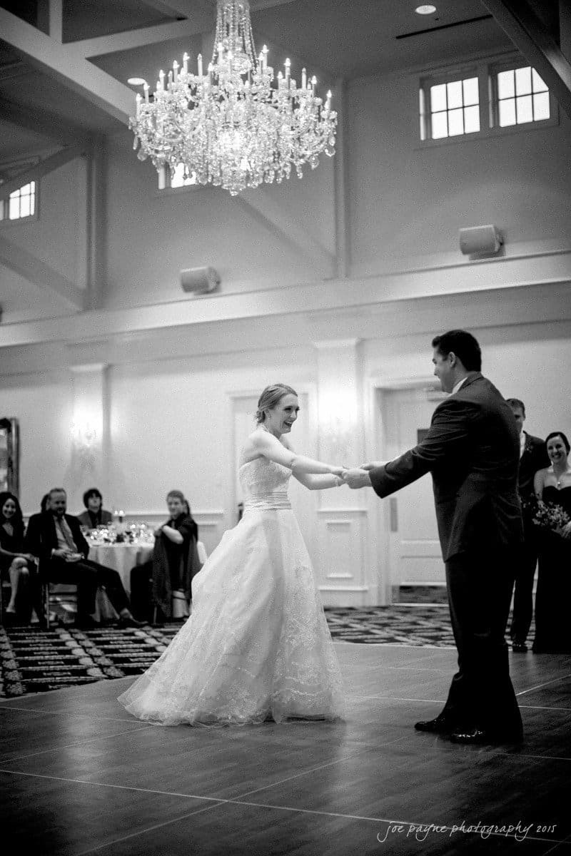 charlotte wedding photographers - ashley & matt A bride and groom share their first dance in a elegant ballroom. The bride wears a white gown, and the groom is in a dark suit. Guests are seated at tables, watching the couple. A large chandelier hangs above them, illuminating the scene.