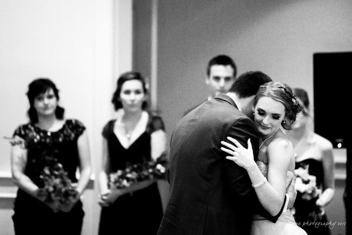charlotte wedding photographers - ashley & matt A couple shares a tender dance at a wedding, with the bride smiling and resting her head on the grooms shoulder. In the background, bridesmaids and groomsmen stand, holding flowers, watching the couple. The image is in black and white.