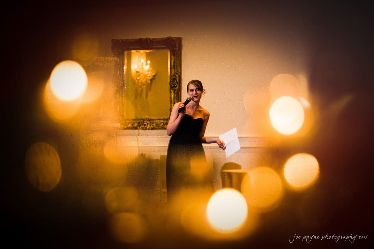 charlotte wedding photographers - ashley & matt A woman in a black dress holds a microphone and a paper, standing in a warmly lit room. Soft golden lights and a framed mirror create a cozy atmosphere.