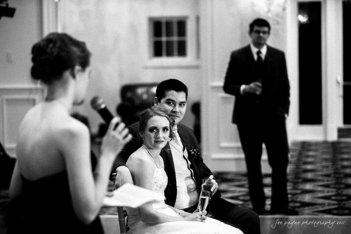 charlotte wedding photographers - ashley & matt A bride and groom sit together, listening to a speech during their wedding reception. The bride holds a champagne glass while a woman speaks into a microphone. A man in a suit stands in the background. The setting is a formal indoor venue.