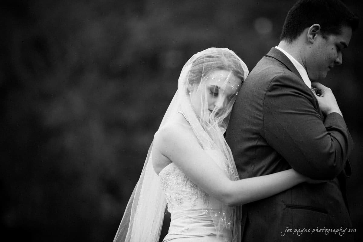 charlotte wedding photographers - ashley & matt A bride in a wedding dress, with a veil partially covering her face, leans against the groom, who is wearing a suit. The couple stands close together, with the brides arm around the groom, in an intimate, black and white portrait.