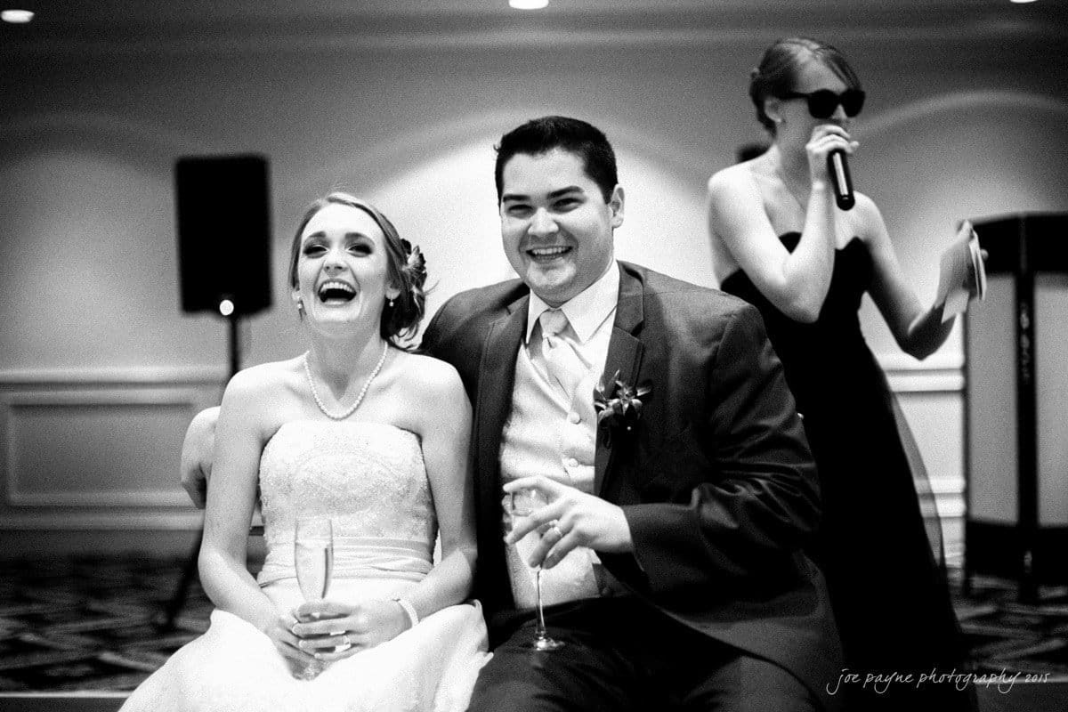 charlotte wedding photographers - ashley & matt A joyful bride and groom sit closely together, both smiling broadly. The bride is in a strapless gown, holding a drink. The groom is in a suit, also holding a drink. A woman in sunglasses and a dress speaks into a microphone in the background.