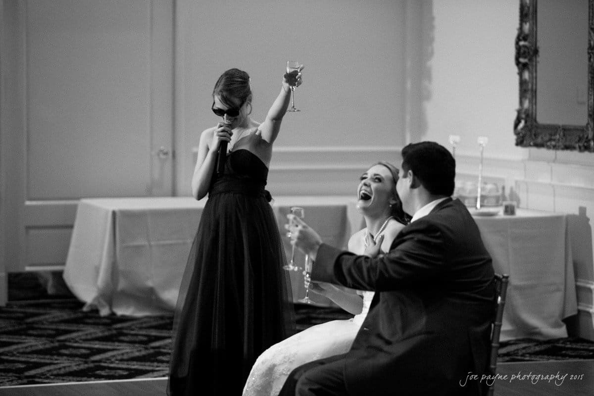 charlotte wedding photographers - ashley & matt A person in a black dress and sunglasses raises a glass while speaking into a microphone. A bride and groom seated nearby laugh and hold their drinks in a celebratory toast in a formal setting.