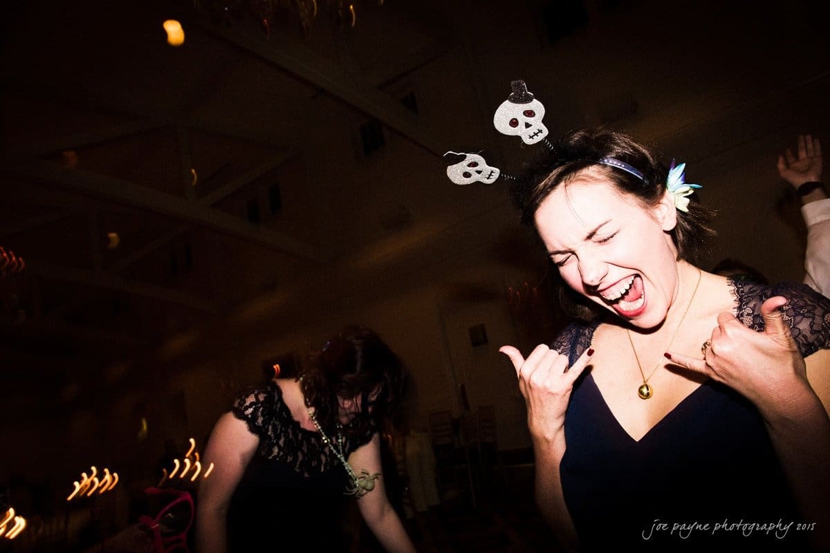 charlotte wedding photographers - ashley & matt A woman with skull hair accessories smiles and makes a rock gesture with her hands at a party. Another person dances in the background. The setting is dimly lit with blurred lights, creating a lively atmosphere.