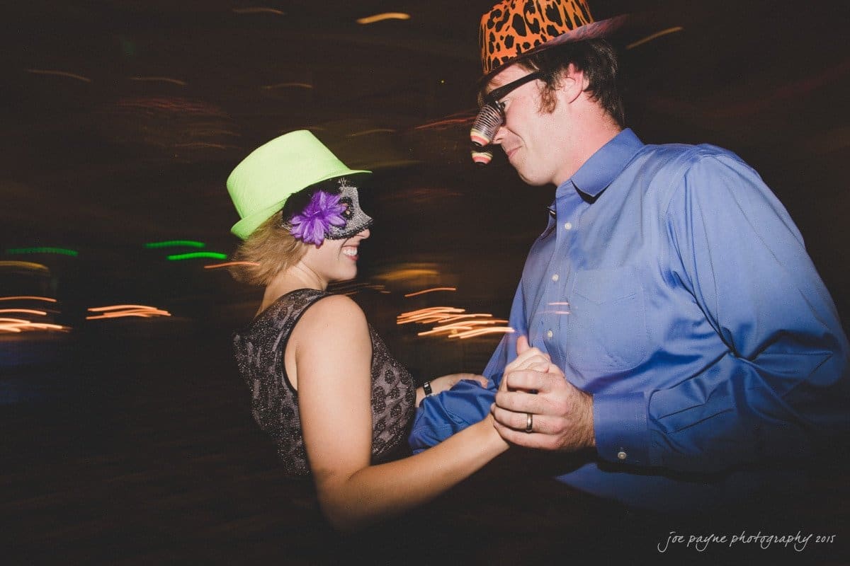 charlotte wedding photographers - ashley & matt A couple in masks and hats dance together. The woman wears a green hat with a purple flower, while the man wears a leopard-print hat and glasses with a fake nose. The setting has dim lighting and blurred background lights.