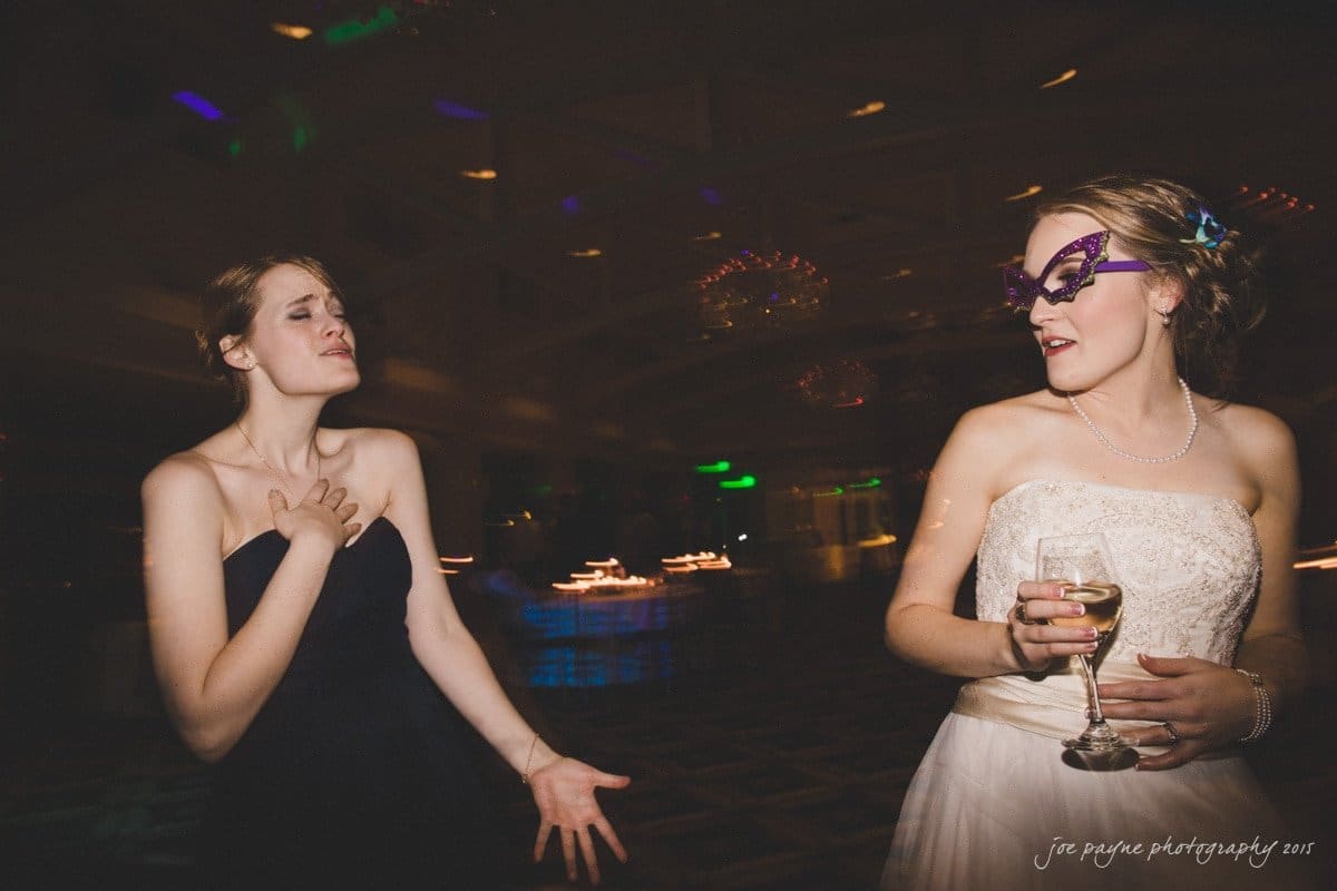 charlotte wedding photographers - ashley & matt Two women are dancing at a formal event. One is wearing a black dress and has her hand on her chest. The other, wearing a white dress and masquerade mask, holds a champagne glass. The background is dimly lit with colorful lights.