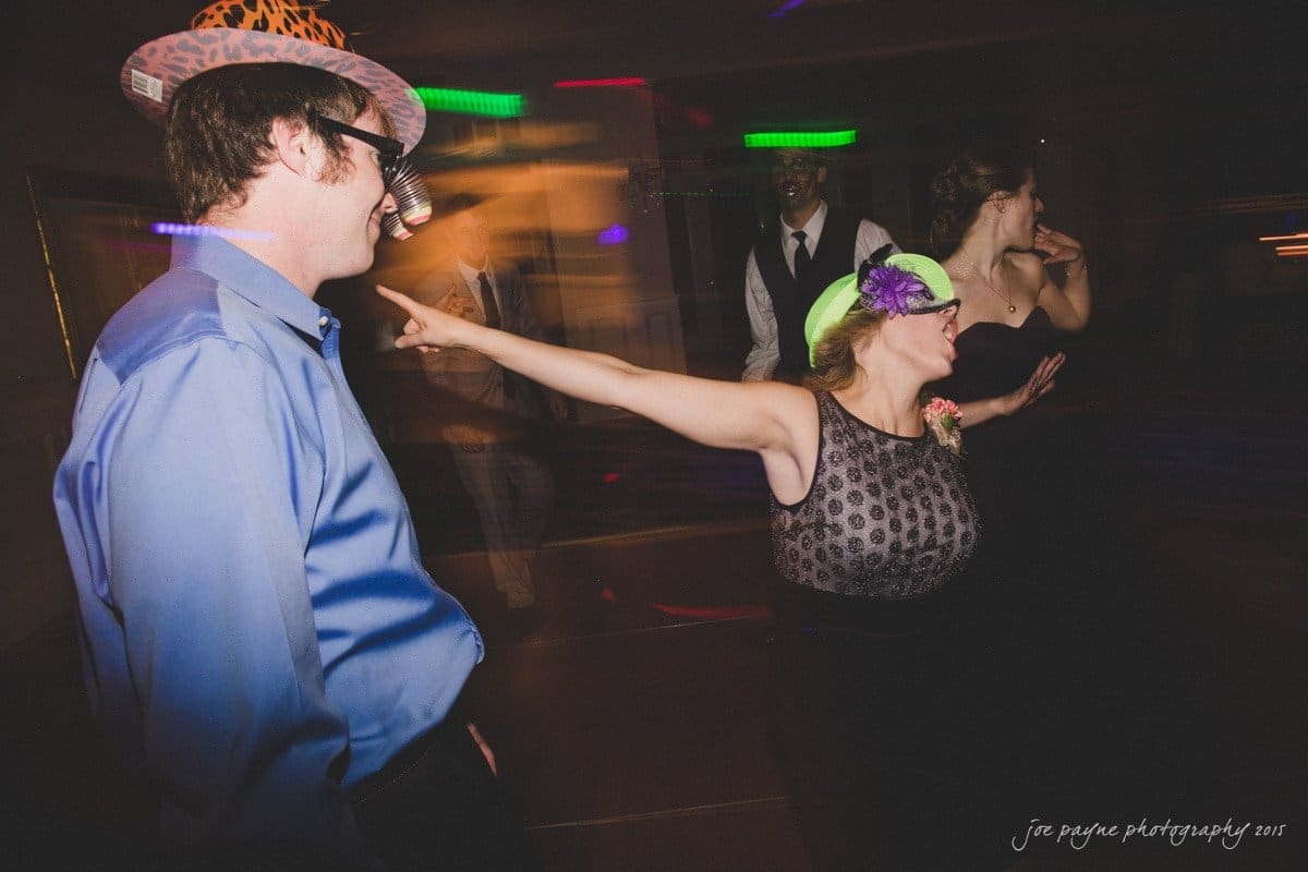 charlotte wedding photographers - ashley & matt A person wearing a hat and a sleeveless top energetically dances, pointing their finger. Others, also wearing hats, dance in the background with colorful lights streaming around them.