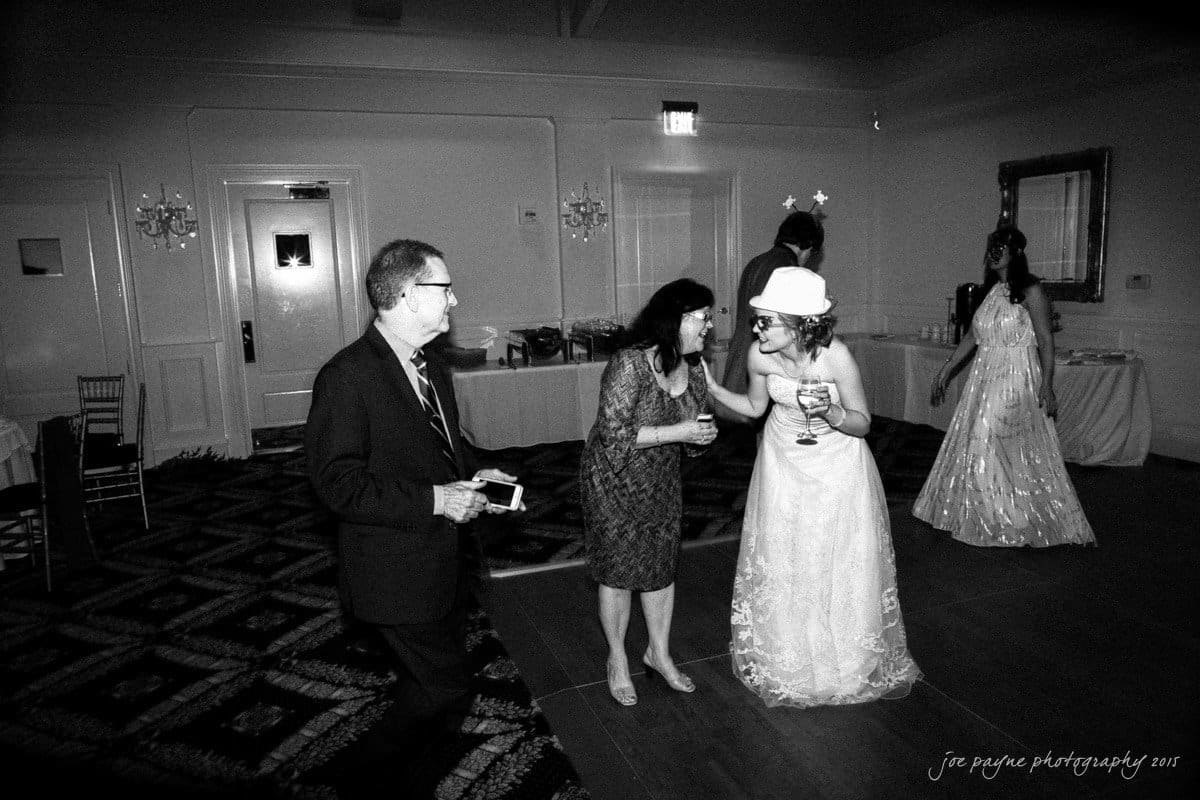 charlotte wedding photographers - ashley & matt A bride in a hat with star-shaped decorations interacts with two guests on a dance floor. The scene is indoors with a patterned carpet and dim lighting. Another woman in a gown stands in the background.