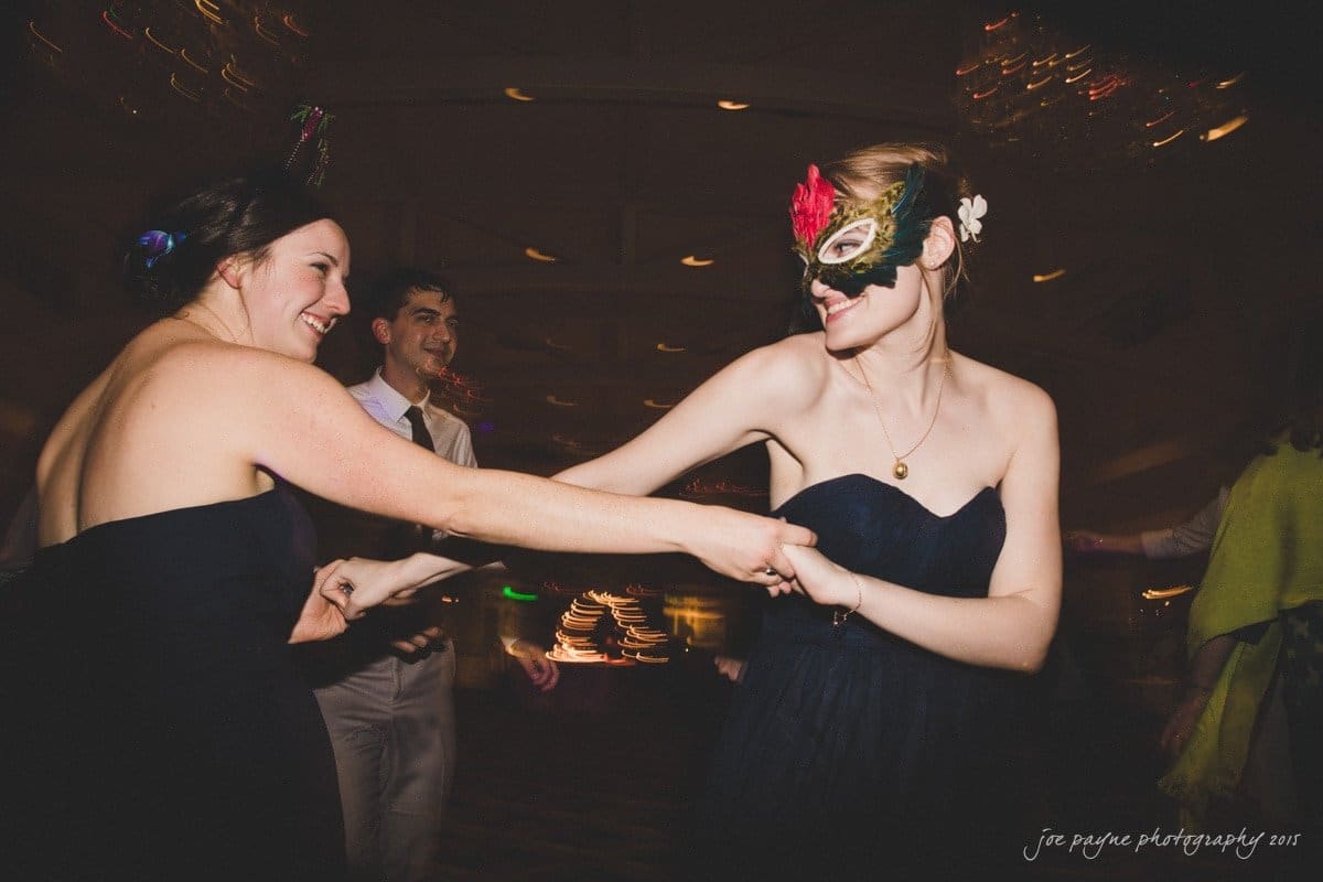 charlotte wedding photographers - ashley & matt Two women in dark dresses dance together at a party. One wears a colorful feathered mask. A man in a suit and a person in a shawl can be seen in the background. The setting has dim, festive lighting.