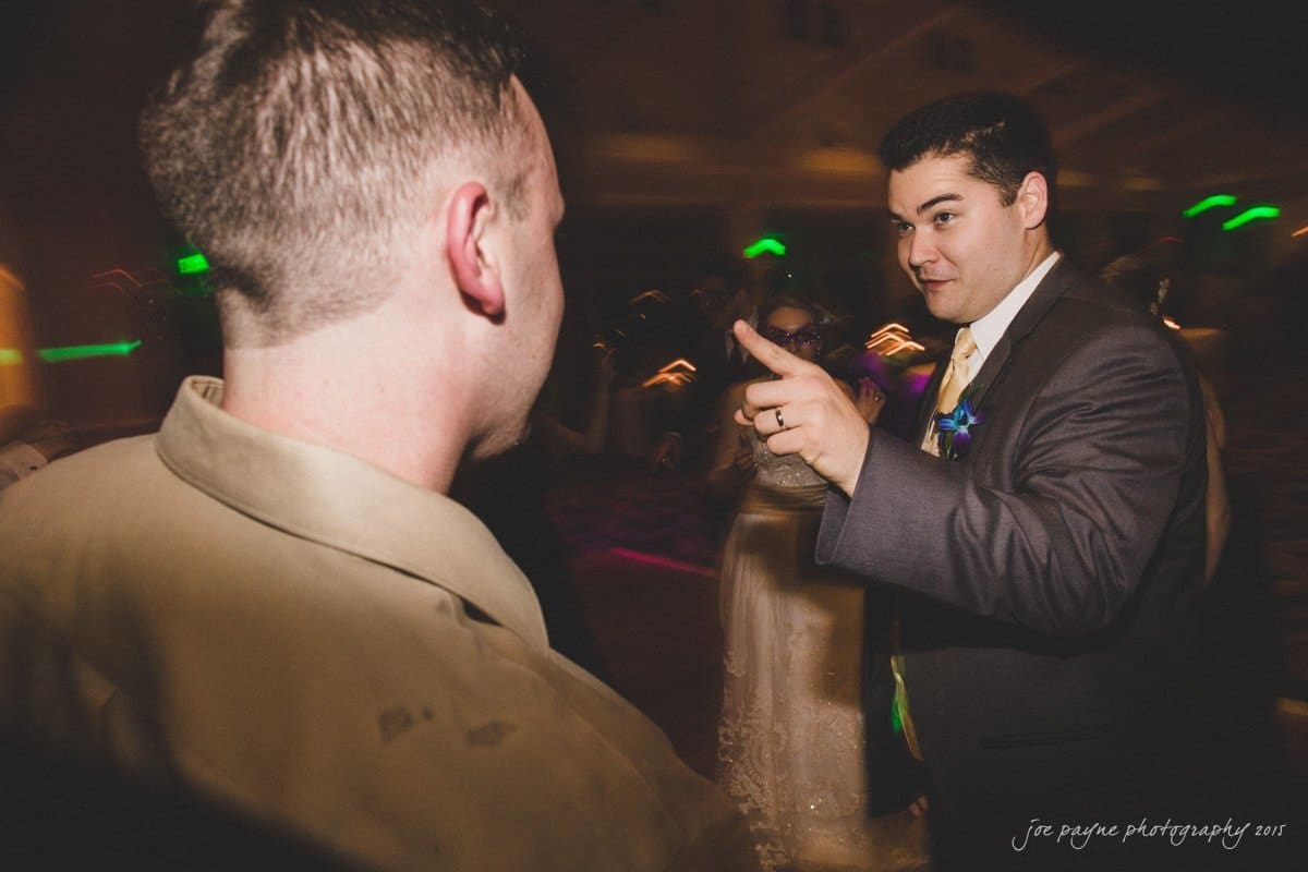 charlotte wedding photographers - ashley & matt Two men are engaged in conversation at an indoor event. One man, wearing a suit and boutonniere, is gesturing with his hand. The lighting is dim with colorful light effects in the background.