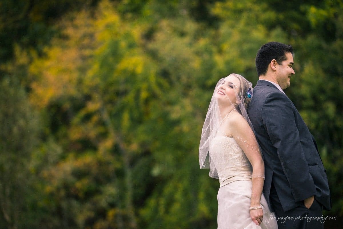 charlotte wedding photographers - ashley & matt Bride and groom stand back-to-back in formal attire, smiling joyfully. The bride wears a veil and strapless gown, while the groom is in a dark suit. They are outdoors with a blurred background of greenery and autumn leaves.