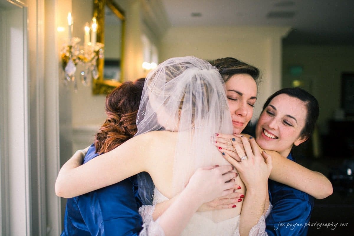 charlotte wedding photographers - ashley & matt A bride in a white dress and veil is embraced by three smiling women in blue robes. The scene is set indoors, with soft lighting from a chandelier and a mirror reflecting part of the room.