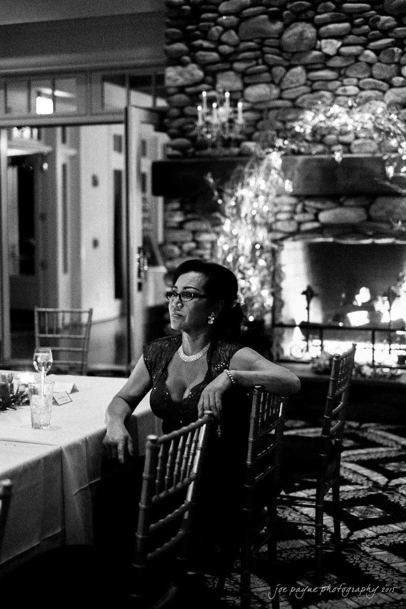charlotte wedding photographers - ashley & matt A woman in a dark dress sits at a table in a dimly lit room with a stone fireplace. She rests her arm on a chair back, surrounded by elegant decor. Reflections of candlelight can be seen in the background.