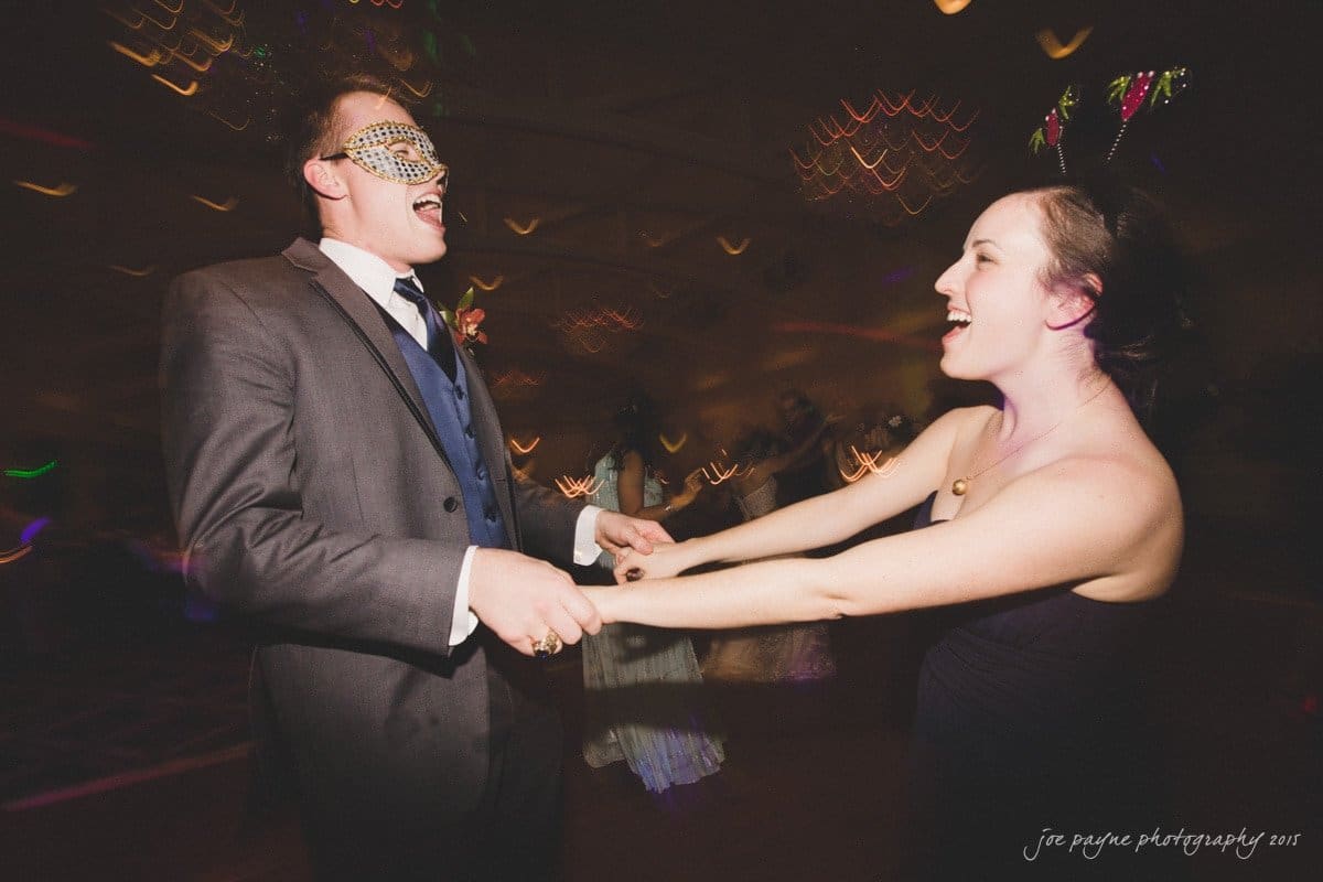 charlotte wedding photographers - ashley & matt A couple dances at a masquerade ball. The man wears a suit and a decorative mask, while the woman is in a black dress with bunny ears. The blurred background features soft lights, creating a festive atmosphere.