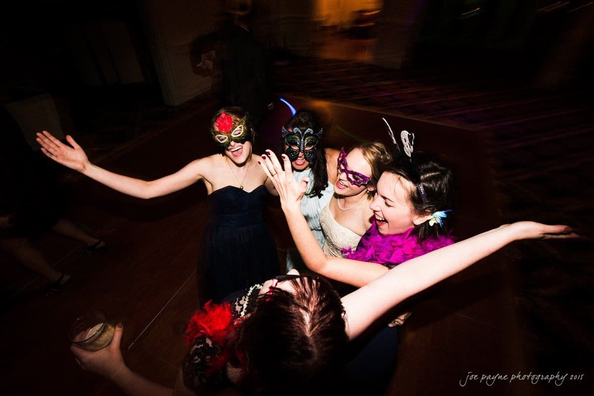 charlotte wedding photographers - ashley & matt Five people in colorful masks and party attire joyfully dance at a masquerade ball. They have their arms raised and are smiling, creating a lively and festive atmosphere. The background is dimly lit, enhancing the vibrant mood.