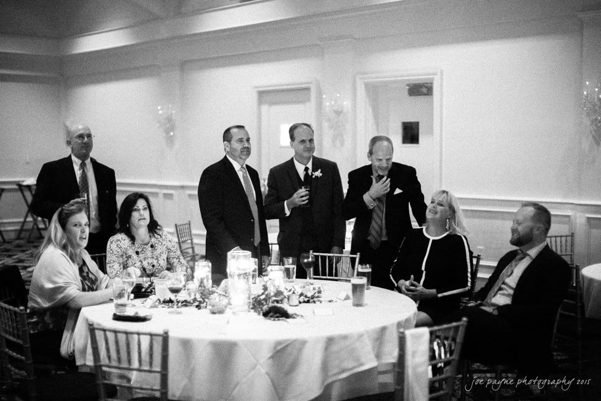charlotte wedding photographers - ashley & matt A group of people in formal attire gathered around a round table with drinks, candles, and decorations. Some are seated, while others stand, engaging in conversation in a warmly lit room.